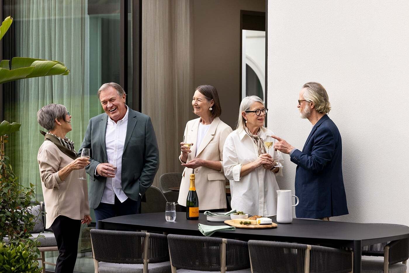 A group of older people standing outside an apartment, laughing and enjoying a drink together.