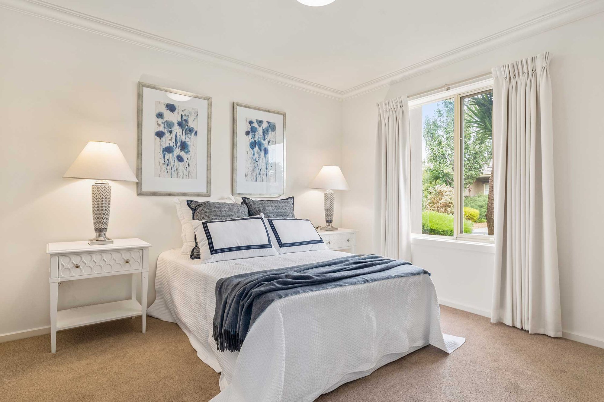 Image of a bedroom with white and blue accents in the linen and hanging pictures and two nightstands with lamps on