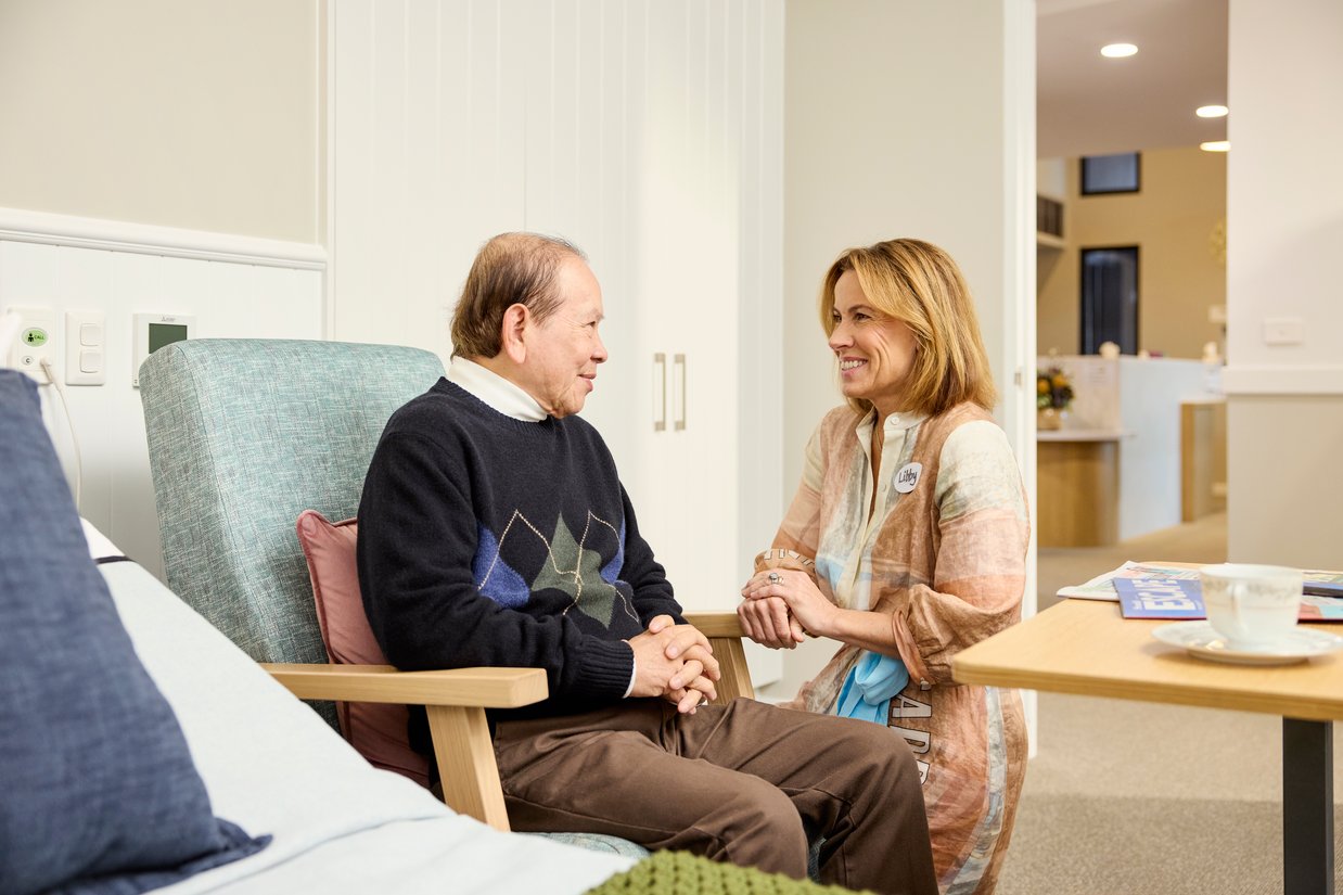 An elderly man sits in a chair, smiling at a carer next to him.